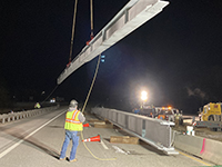 Crane lifts beam into the air as workers supervise below.